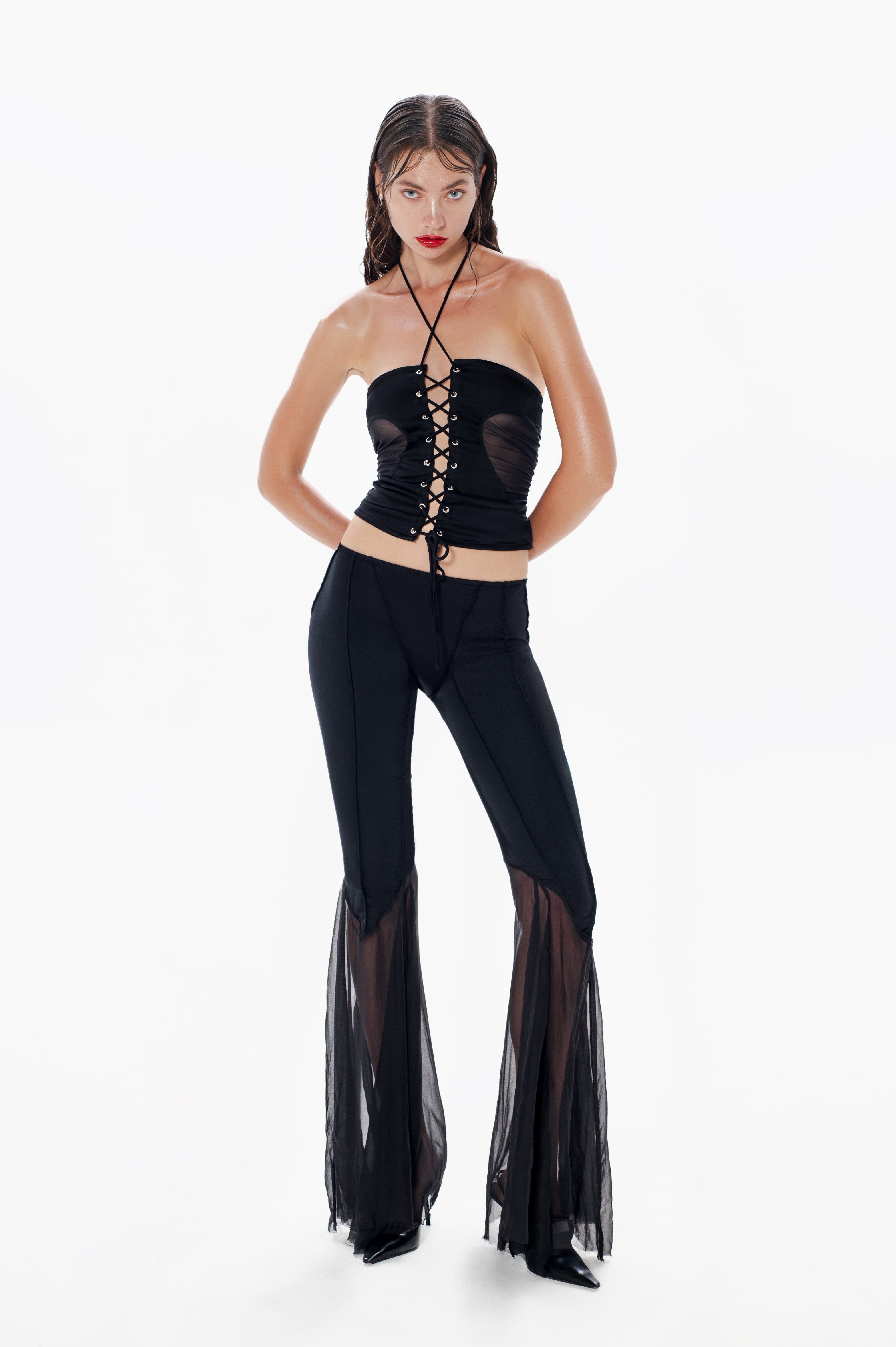 A woman modeling black flare pants with sheer chiffon panels and a contoured, structured lycra body. The outfit includes a front zipper and a pull-on elastic waistband.