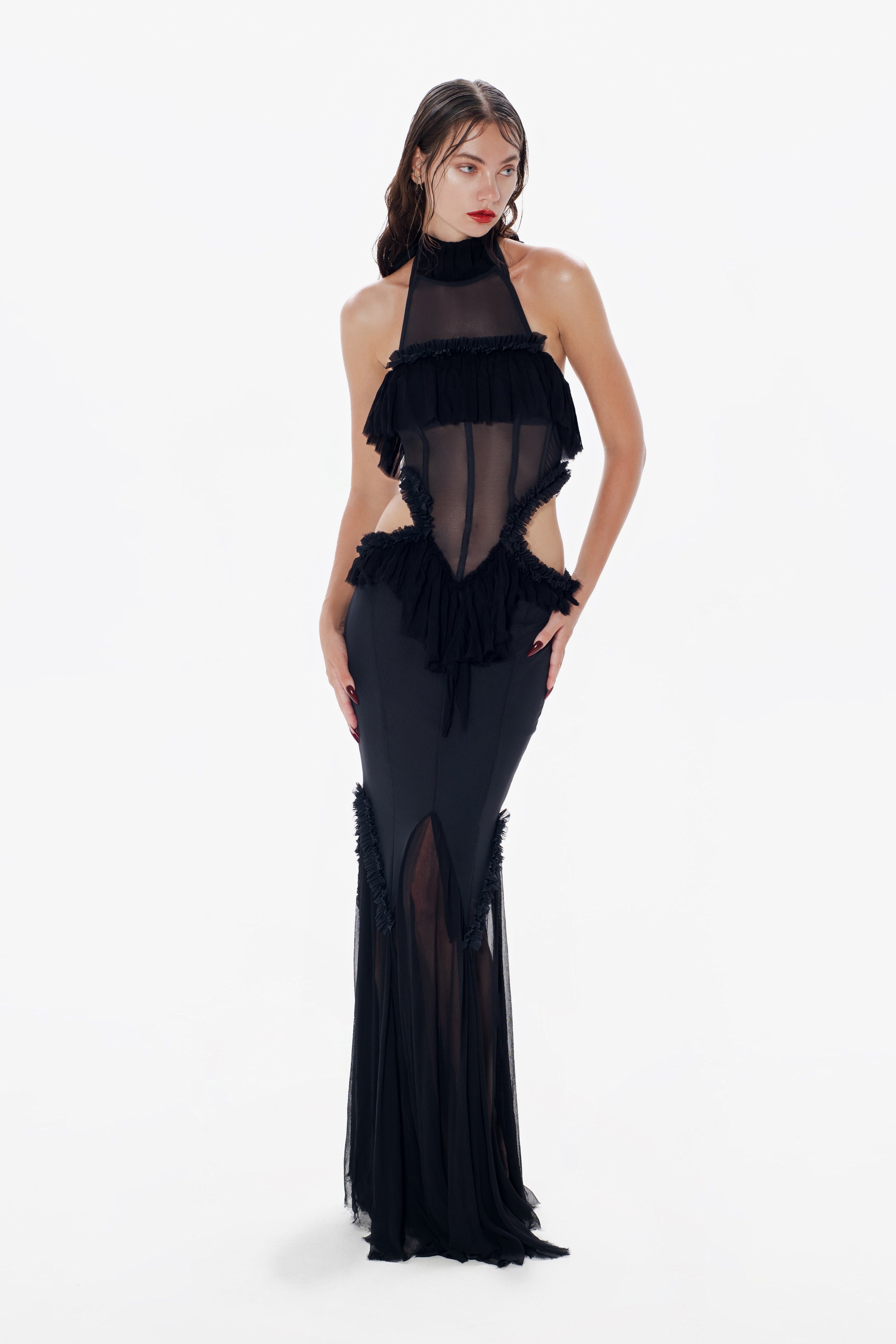 A model wearing a black maxi dress with a halter neckline and mermaid silhouette, featuring cut-outs at the waist and ruffle details.
