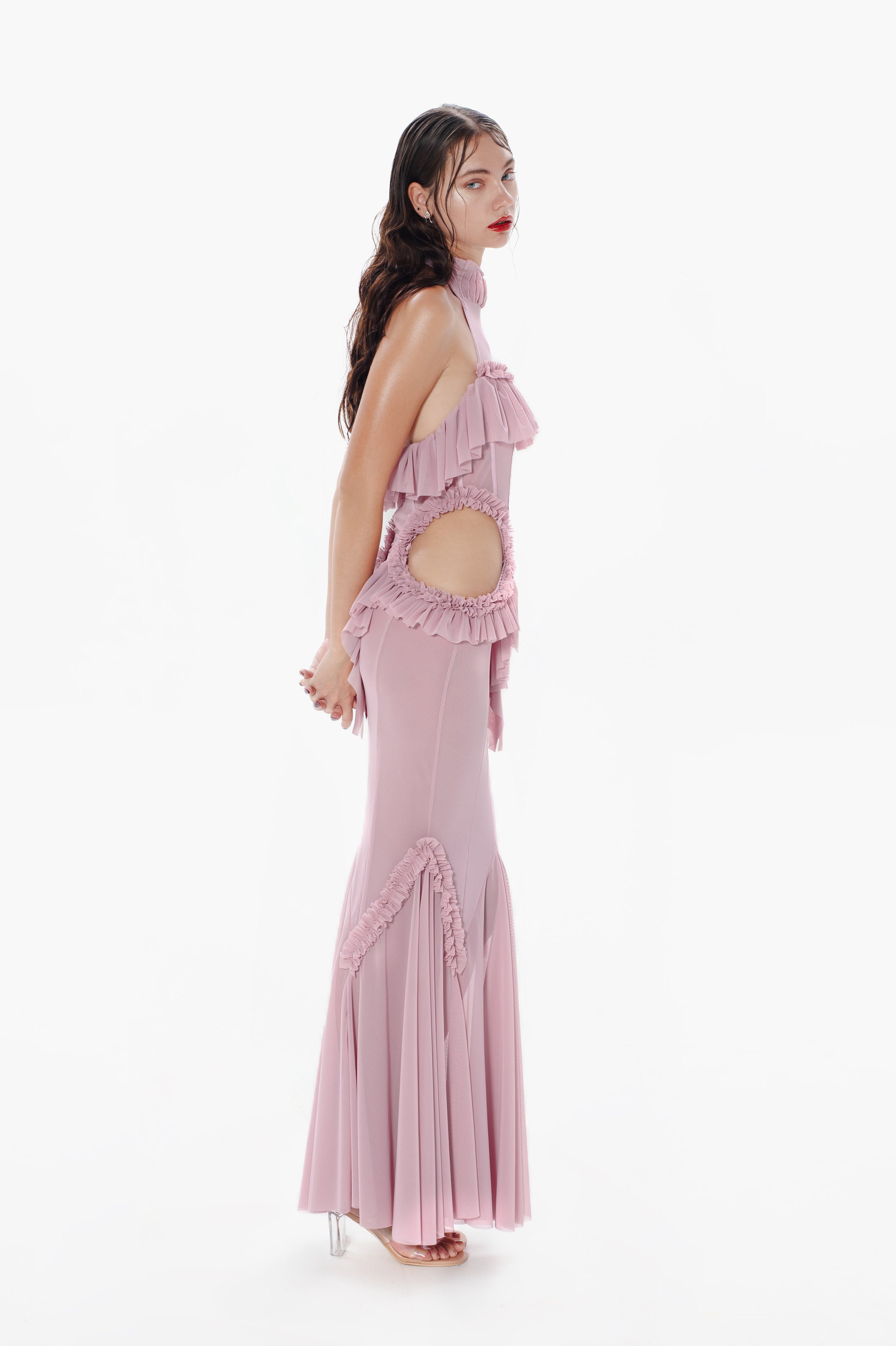 A model wearing a pink maxi dress with a halter neckline and mermaid silhouette, featuring cut-out waist areas and ruffle accents.