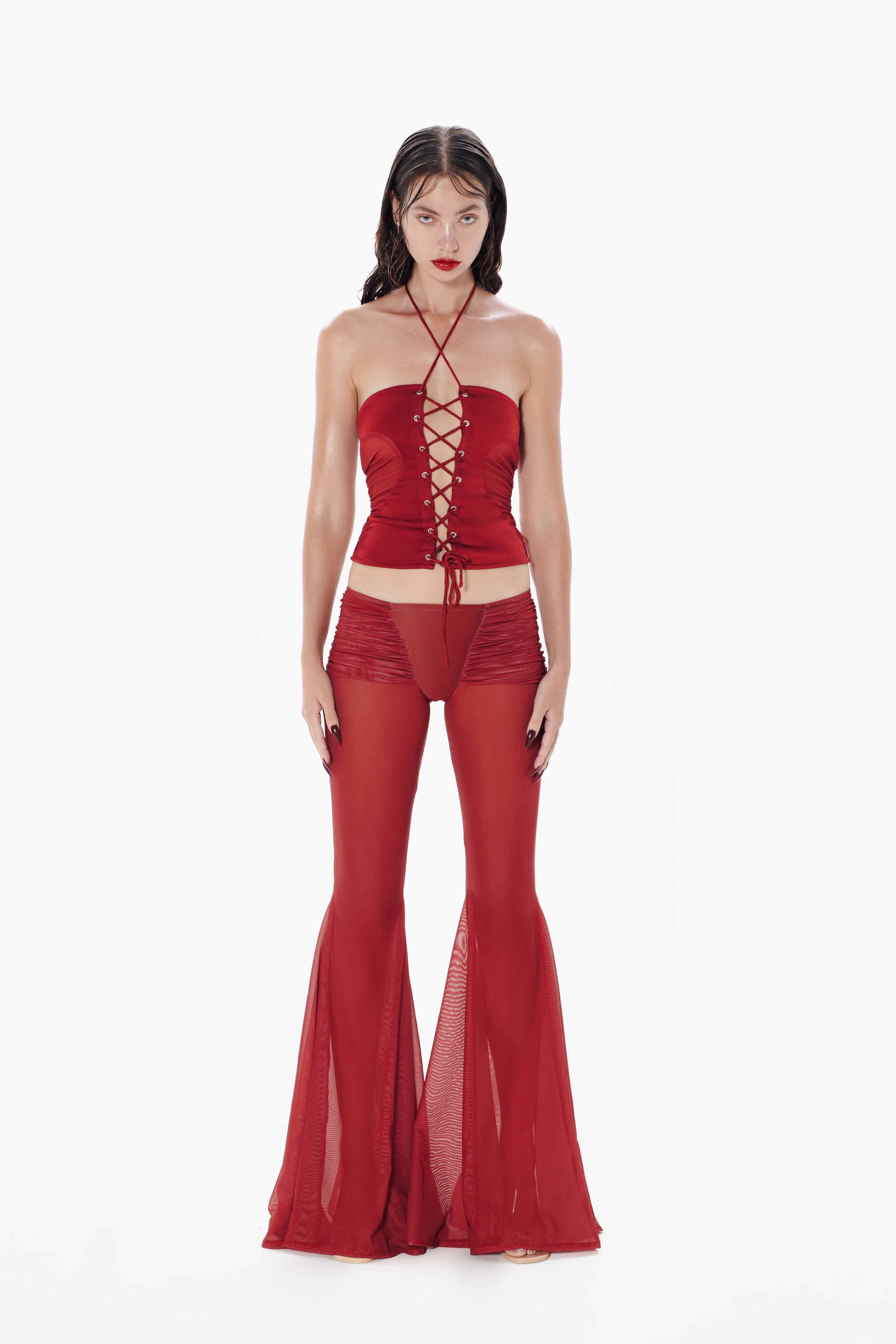 A model wearing red low-rise sheer mesh pants with draping at the front and back, featuring back ruching detail and dramatic flared legs.